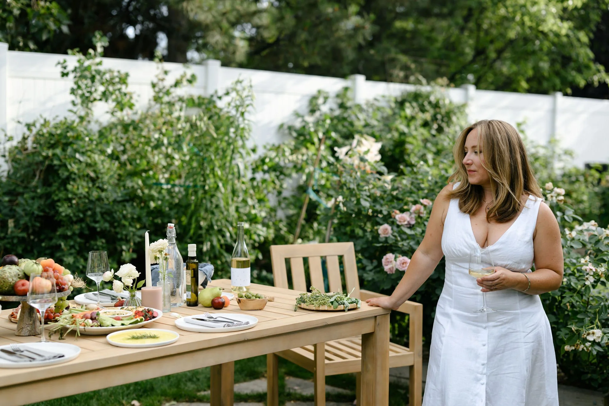 Woman in white dress standing next to a dinner table with food and wine in a garden with green foliage and a white fence in the background.
