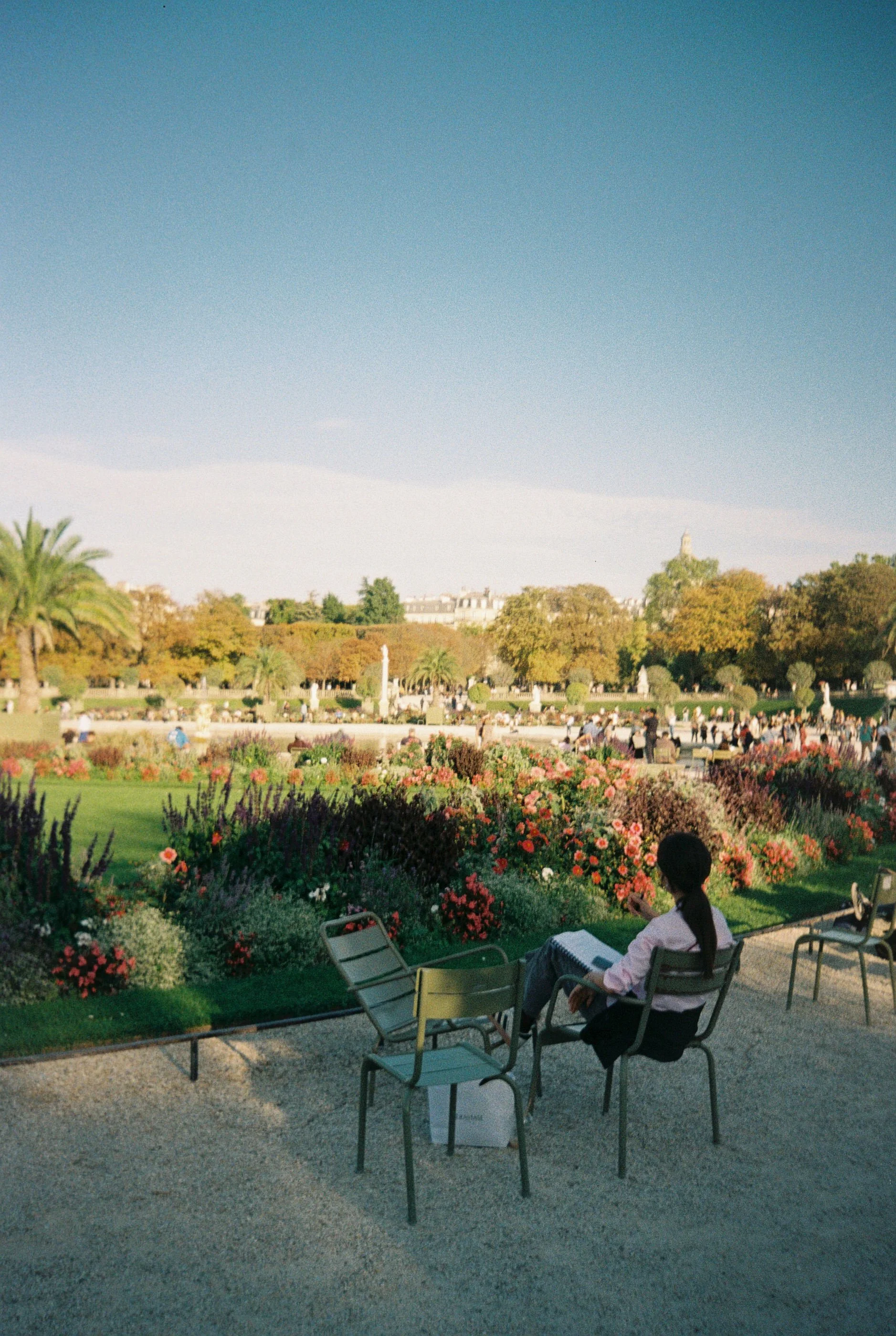 A woman sitting on a chair in a garden with colorful flowers, trees, and a large crowd in the background under a clear blue sky.