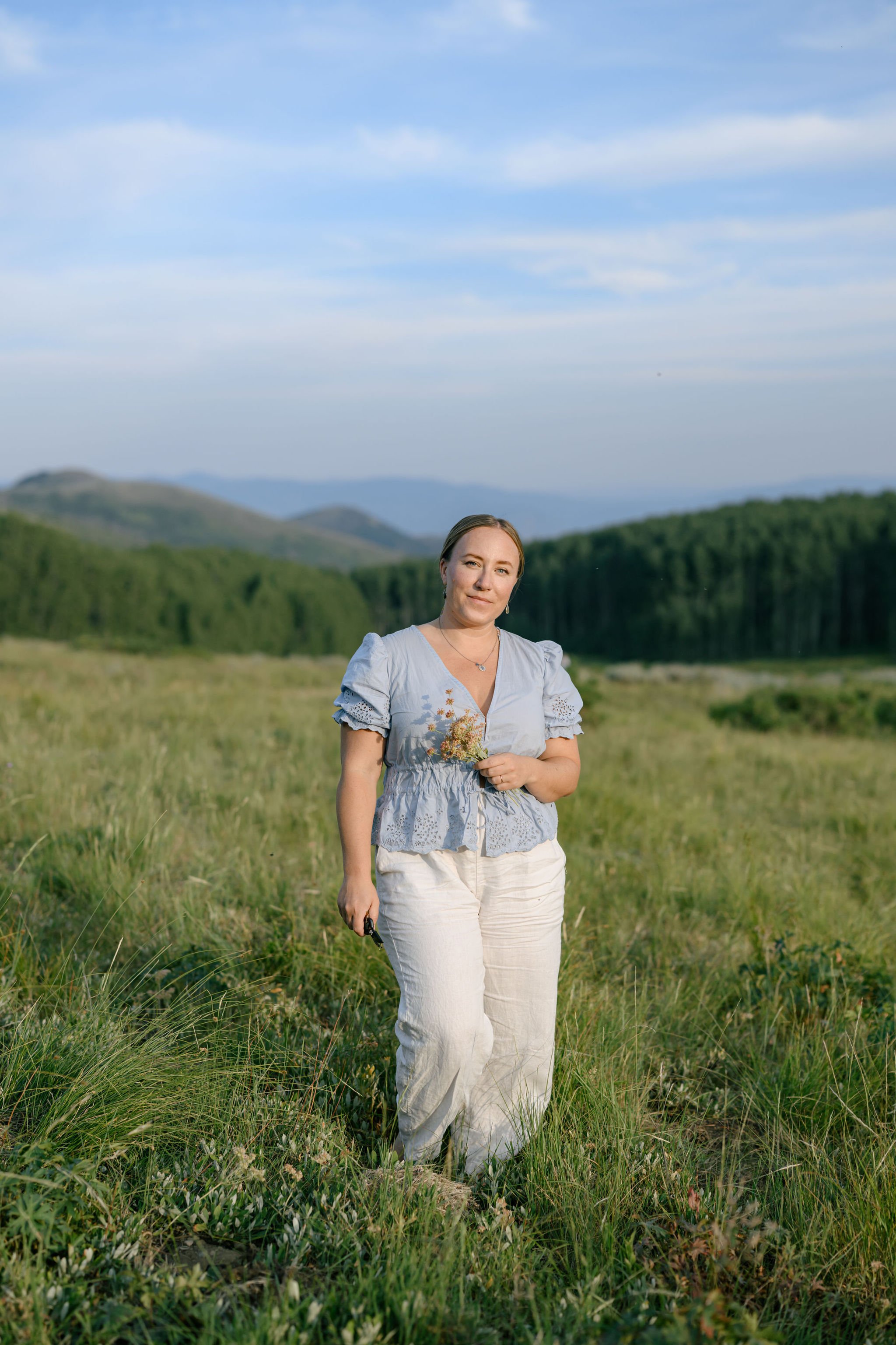 A woman standing in a grassy field holding a small bouquet of flowers, with green hills and a blue sky in the background.